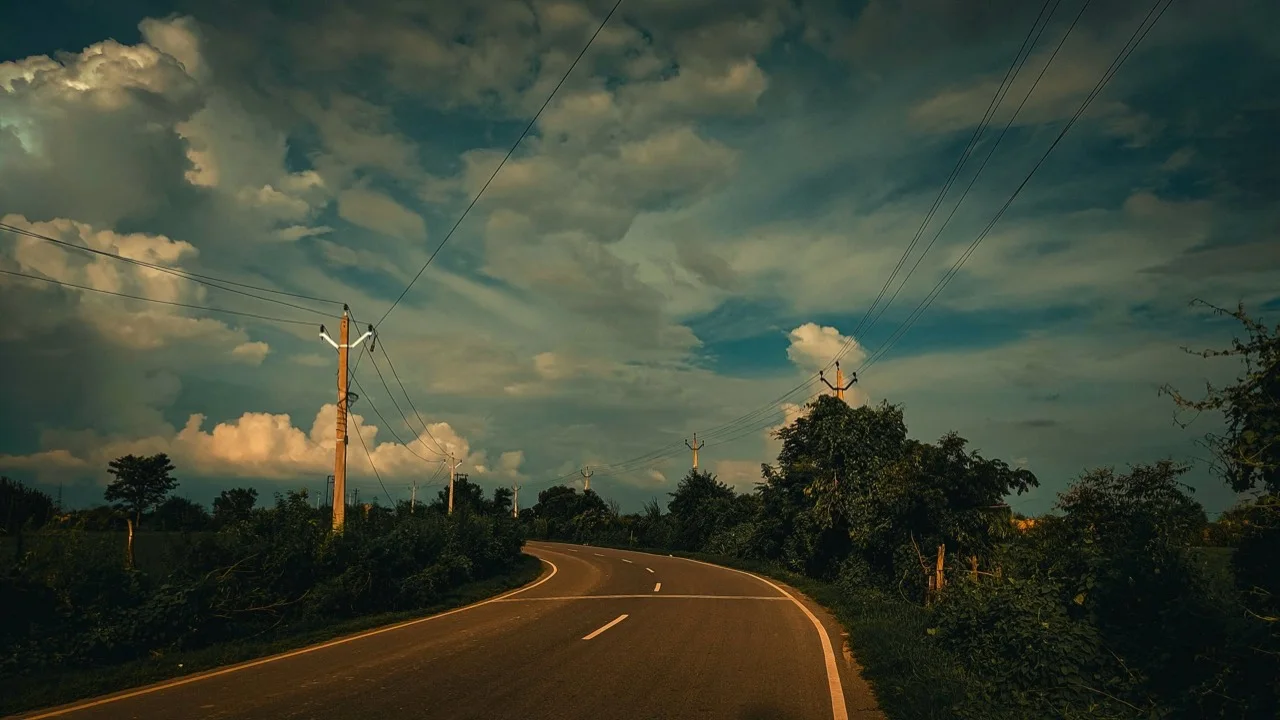 Eine kurvige Straße zwischen grünen Bäumen unter Wolken und Stromleitungen im warmen Licht
